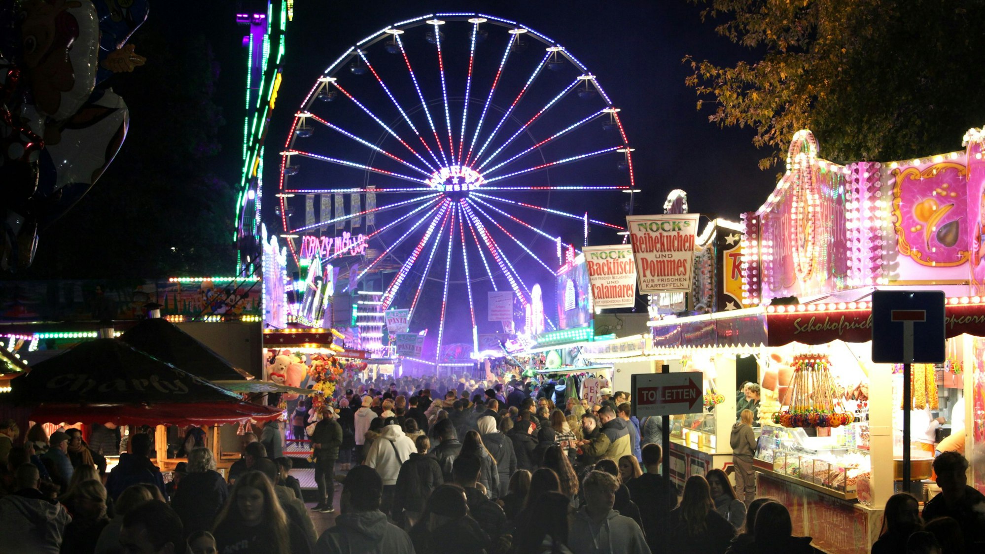 Besucher drängen sich auf einem festlich beleuchteten Jahrmarkt mit Riesenrad und Schaubuden.