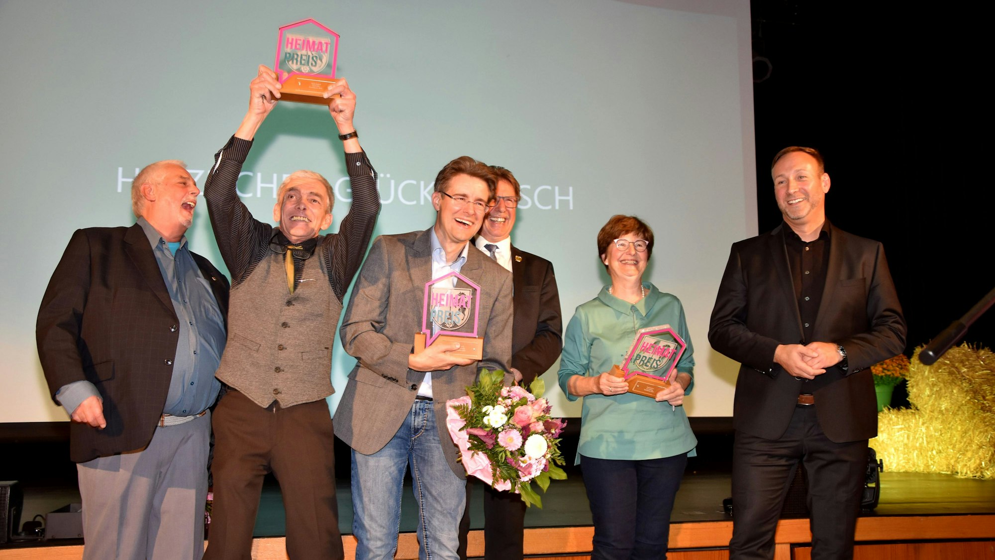 Fünf Männer und eine Frau lachen herzlich, einer der Männer reckt eine Trophäe in die Luft.