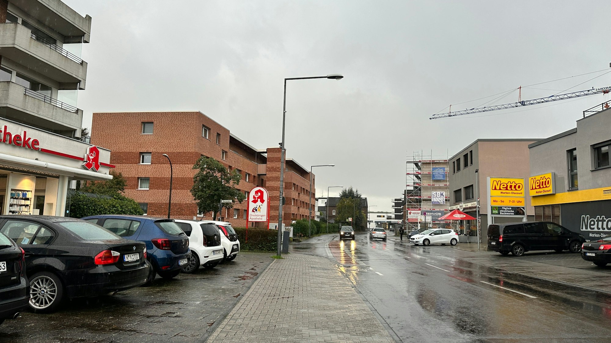 Autos parken vor einem Supermarkt und einer Apotheke, die gegenüber an einer Straße liegen.