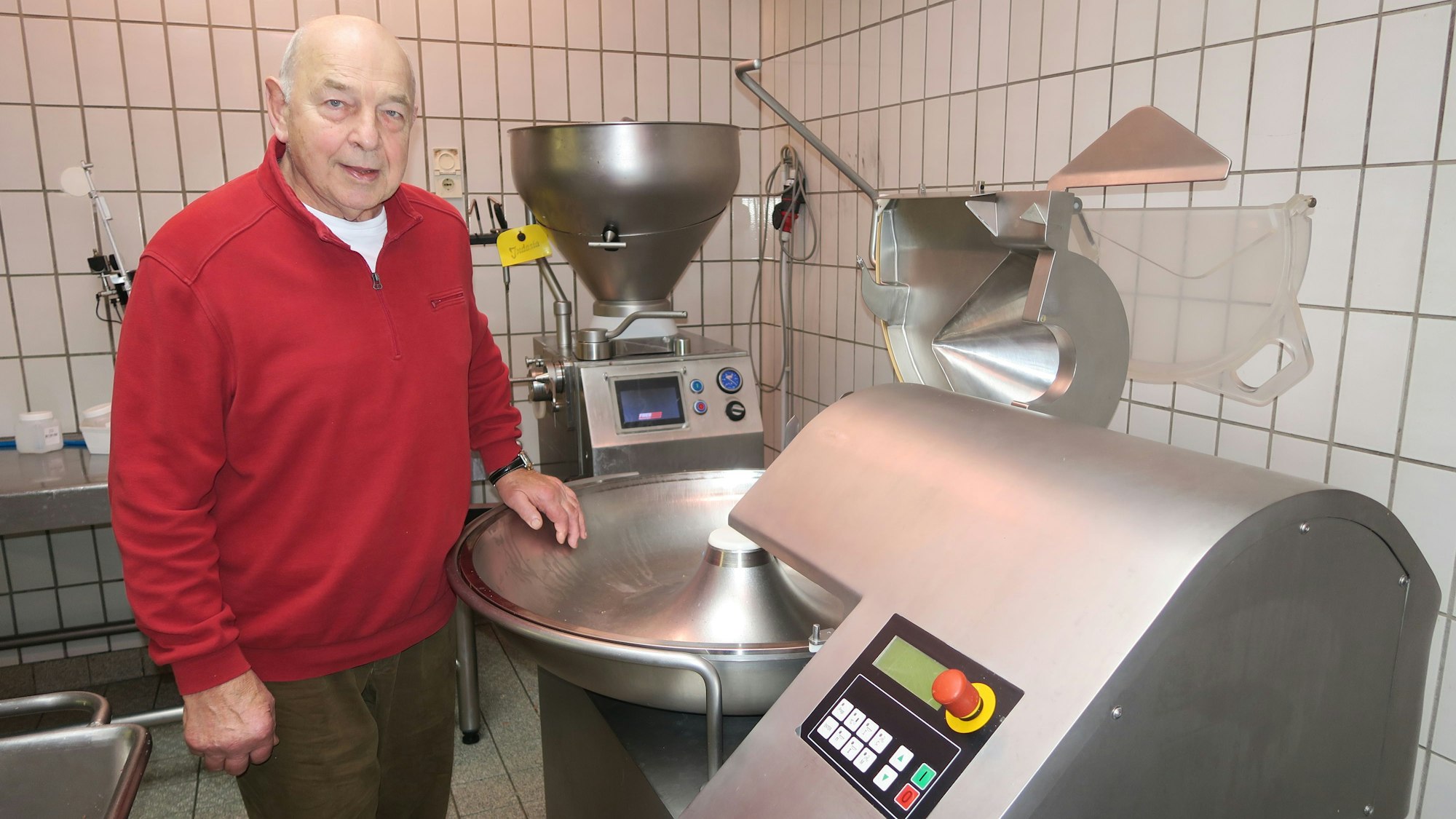 Ein alter Herr im roten Pullover steht neben einer Maschine aus Edelstahl.