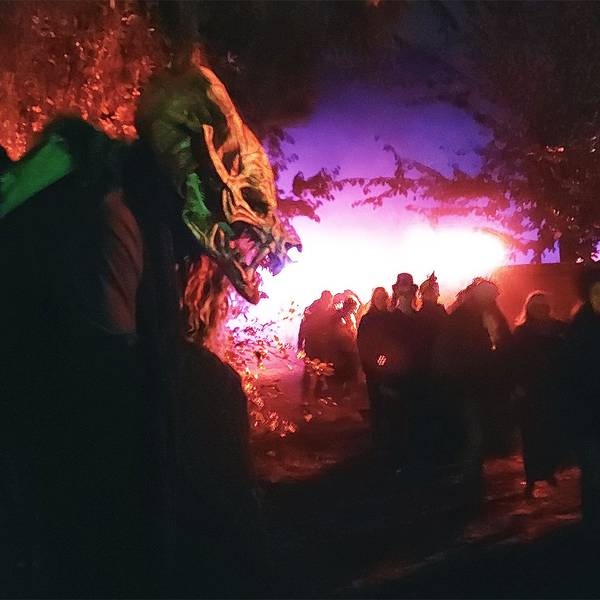 Vorne lauert das maskierte Ungeheuer, während eine Besucherschar vorbeispaziert. Die Szenerie ist in rotes und blaues Licht getaucht.