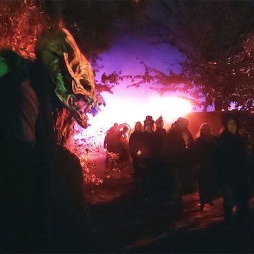 Ein Mensch mit einer gruseligen Monstermasse steht im Dunklen neben einem Weg, auf dem viele andere Menschen laufen. Die Szene ist in rot-violettes Licht getaucht.