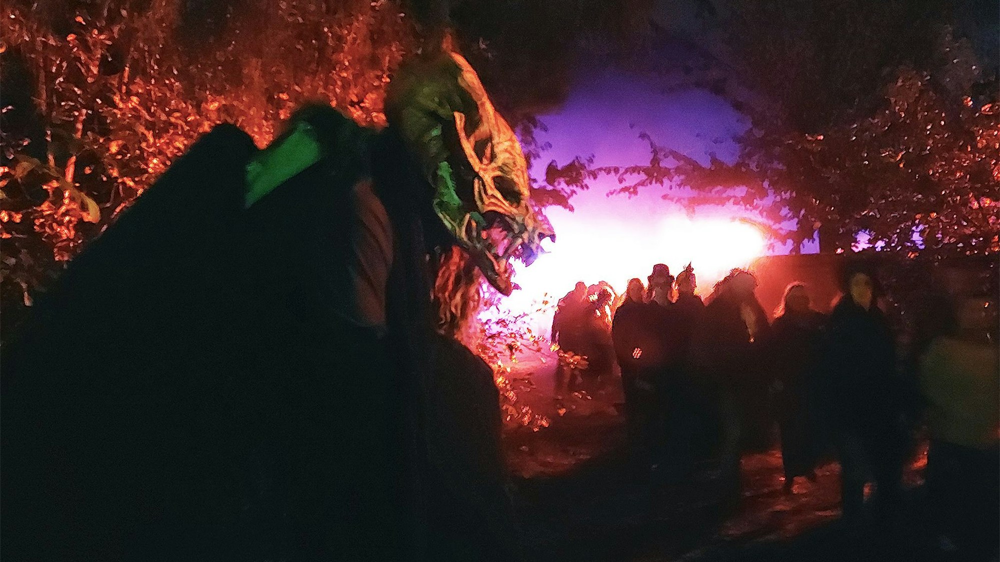 Ein Mensch mit einer gruseligen Monstermasse steht im Dunklen neben einem Weg, auf dem viele andere Menschen laufen. Die Szene ist in rot-violettes Licht getaucht.