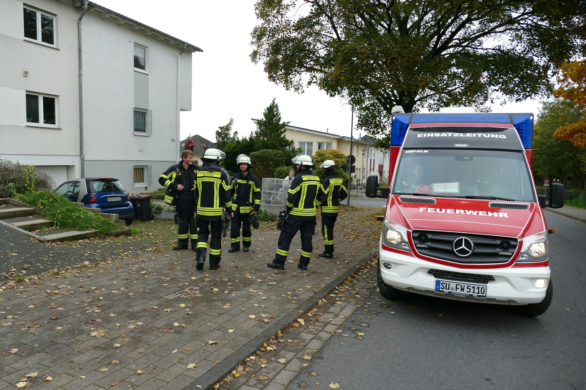 Für Feuerwehrmänner in schwarz-neongelben Uniformen und mit weißen Helmen stehen auf dem Trottoir zwischen einen Feuerwehrfahrzeug und dem betroffenen Wohnhaus.