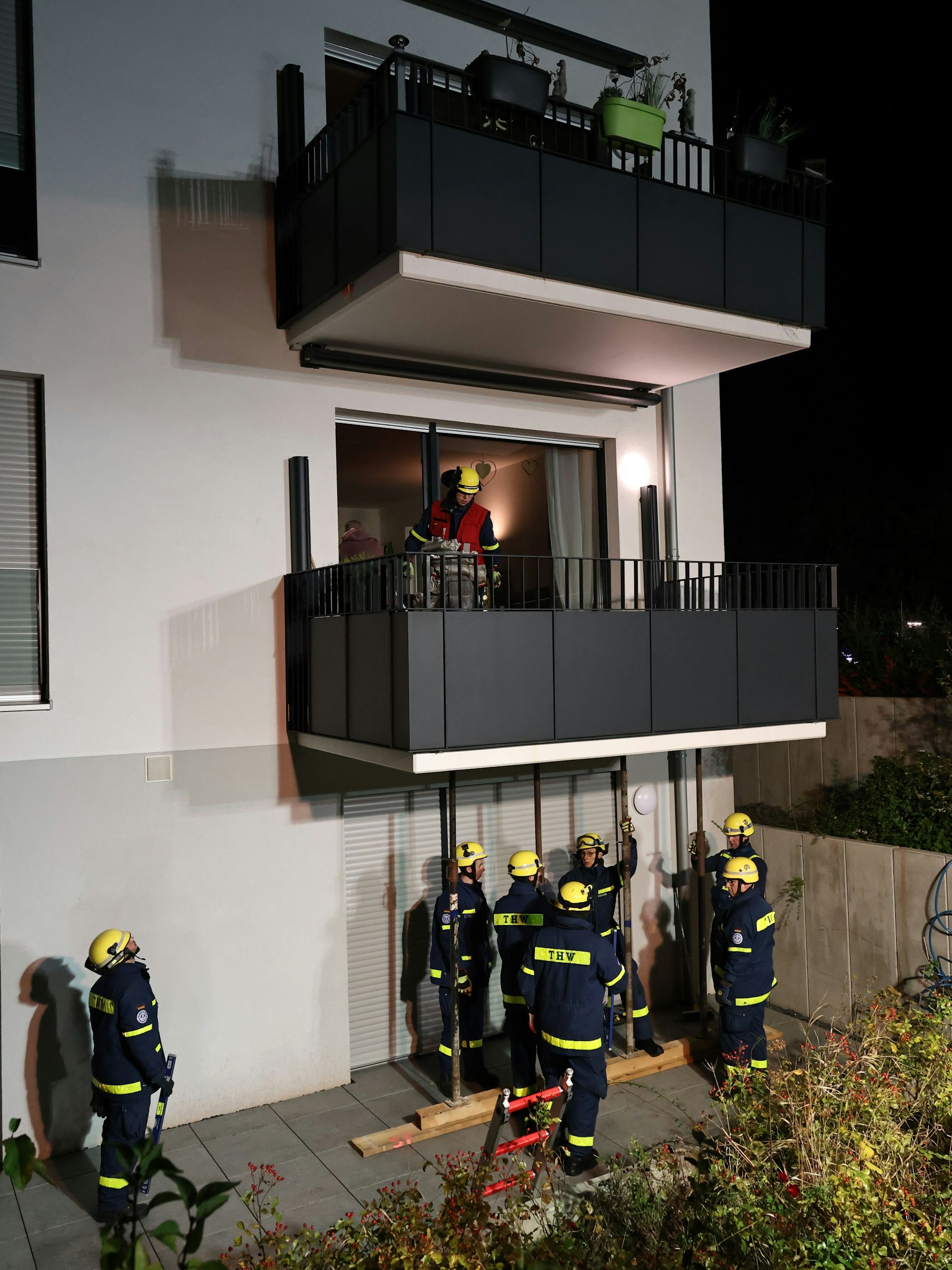 Unten, auf Kellerniveau, sichern THW-Helfer in ihren blauen Monturen und gelben Helmen zunächst den ersten Balkon. Ein Gruppenführer verfolgt die Arbeiten vom ersten Balkon aus, darüber der Balkon, der abzubrechen droht.