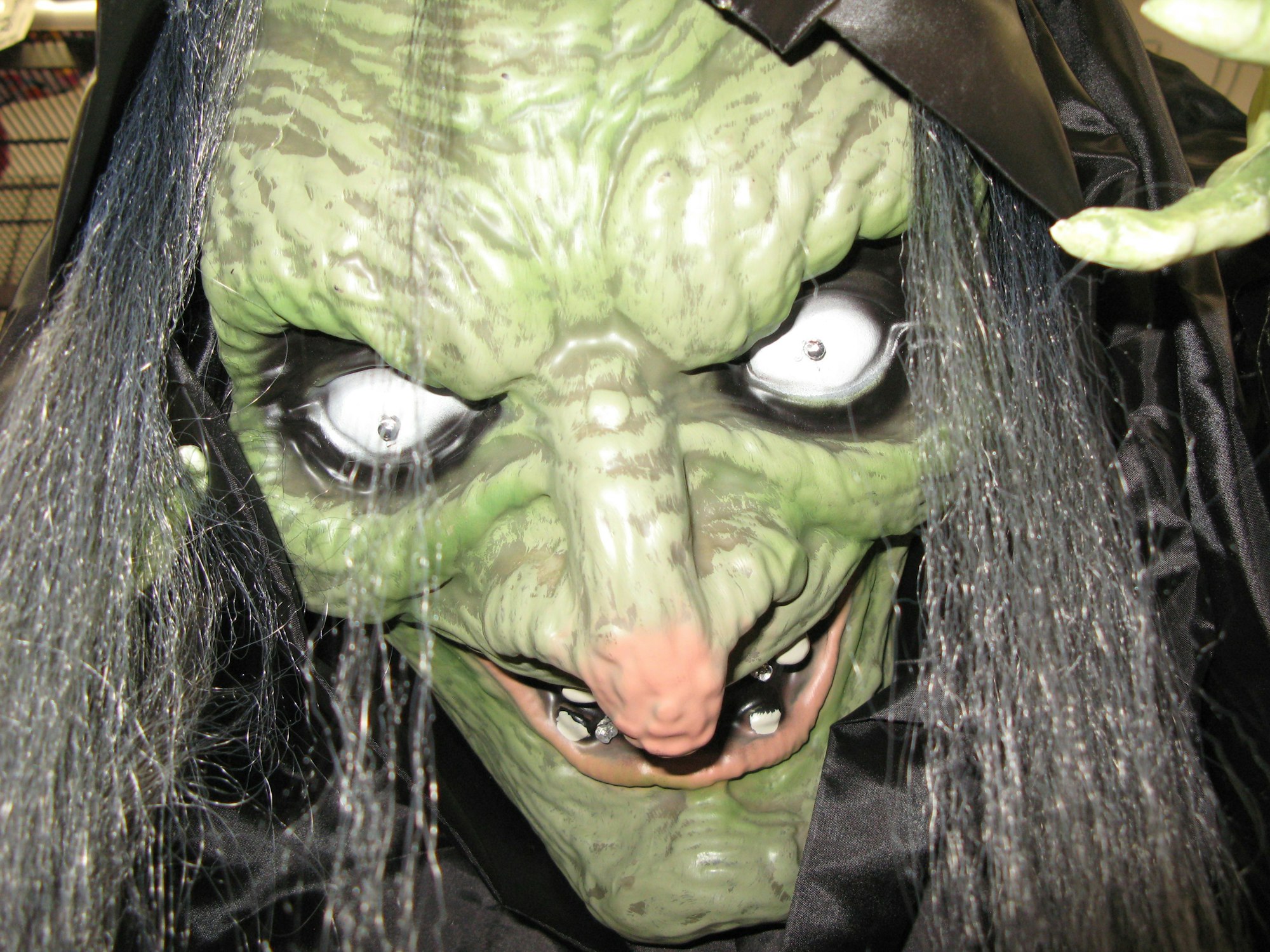 Das Bild zeigt eine Hexenmaske mit grüner Haut, aufgerissenen Augen und langen grau-schwarzen Haaren.
