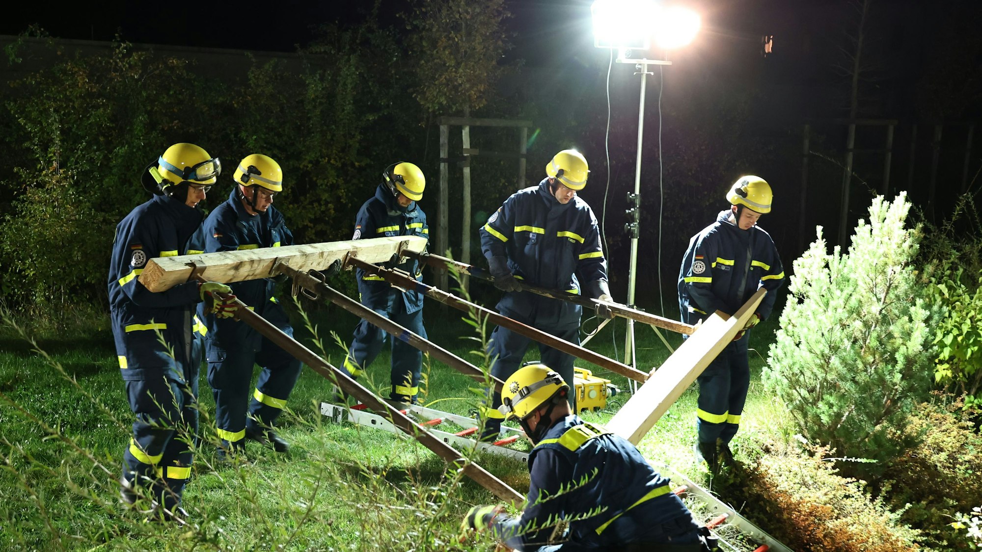 Sechs THW-Helfer tragen die Konstruktion im Licht eines aufgestellten Scheinwerfers die Böschung am Haus hinab.