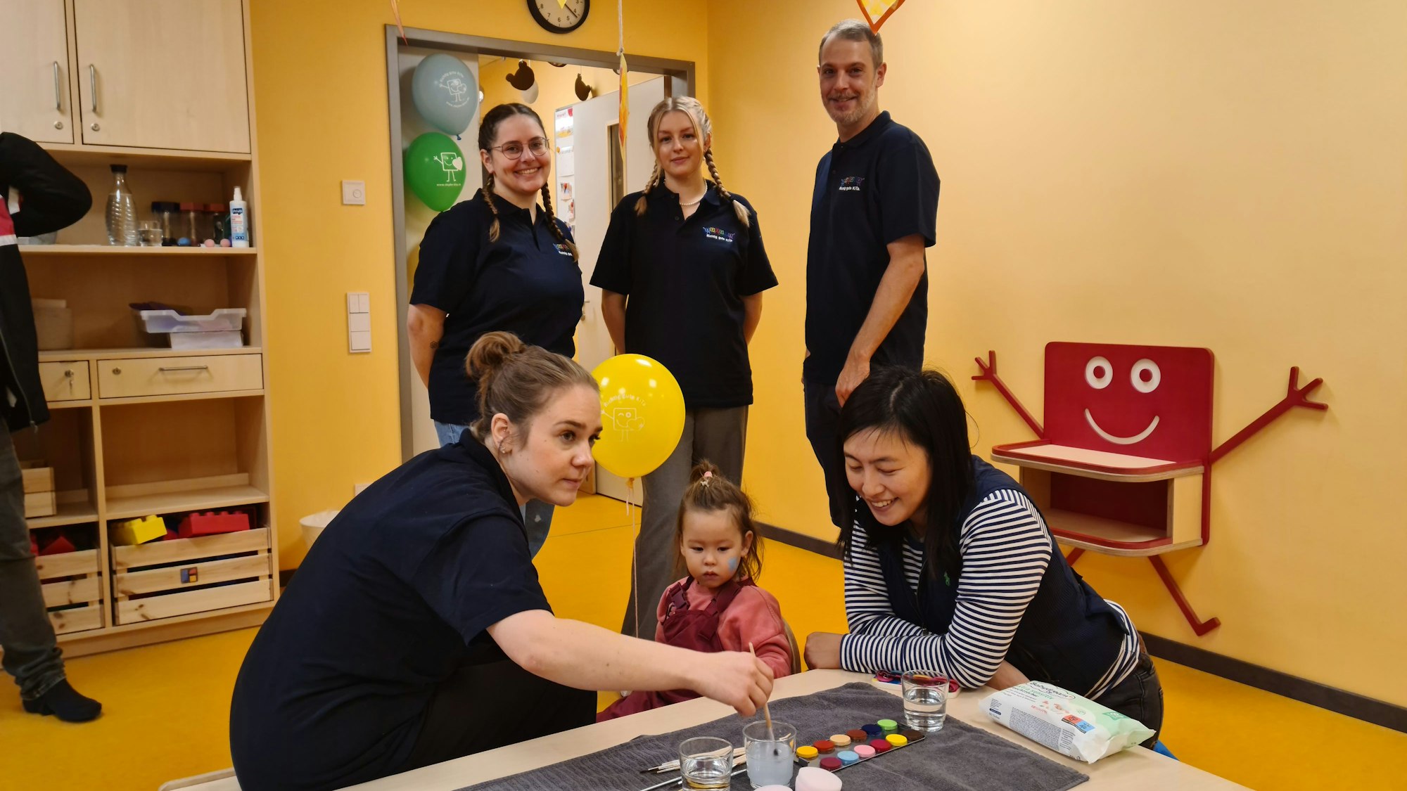 Zwei Frauen und ein Mann stehen hinter zwei Frauen, die mit einem kleinen Mädchen an einem niedrigen Tisch sitzen.
