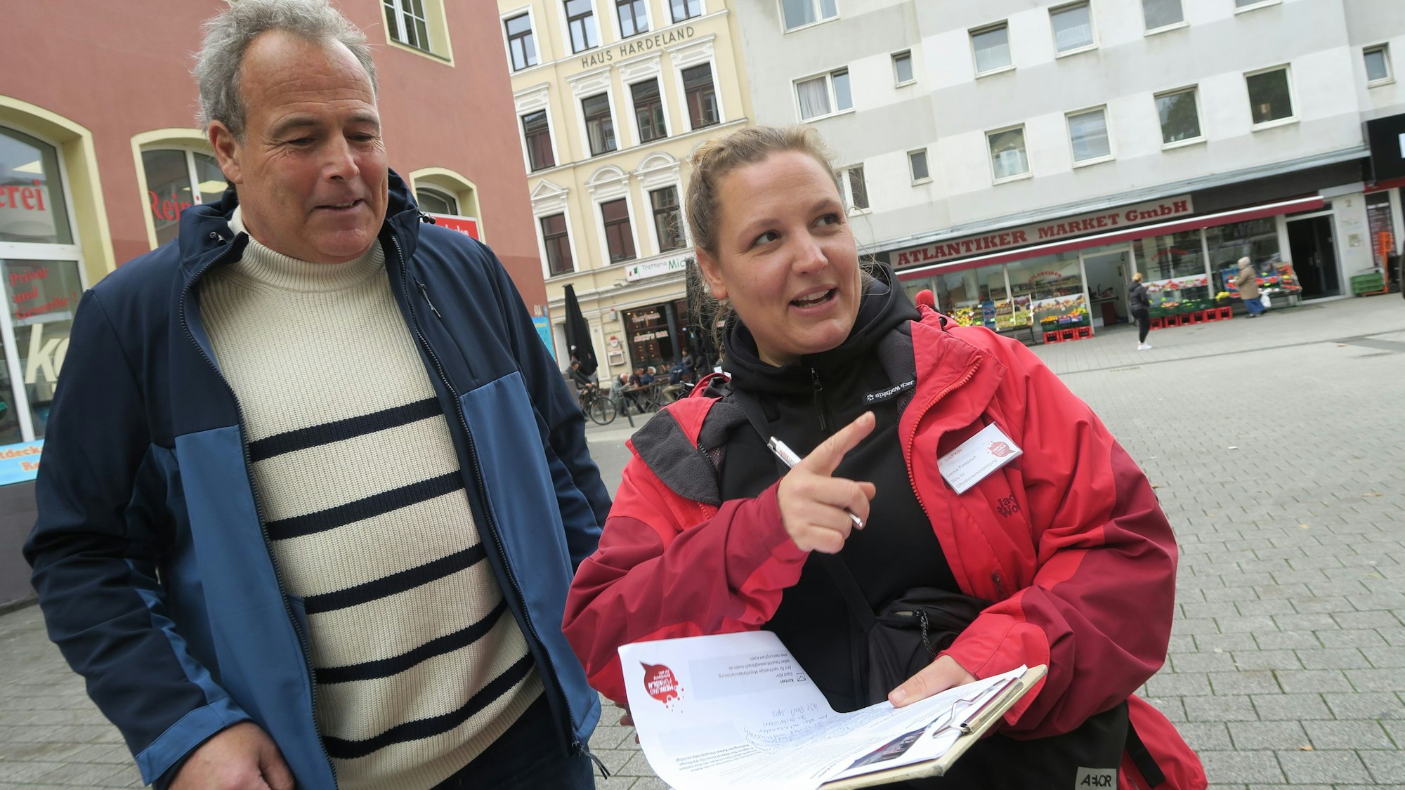 Ein Mann und eine Frau stehen auf einem Platz, die Frau hat ein Clipboard mit einem Fragebogen in der Hand.