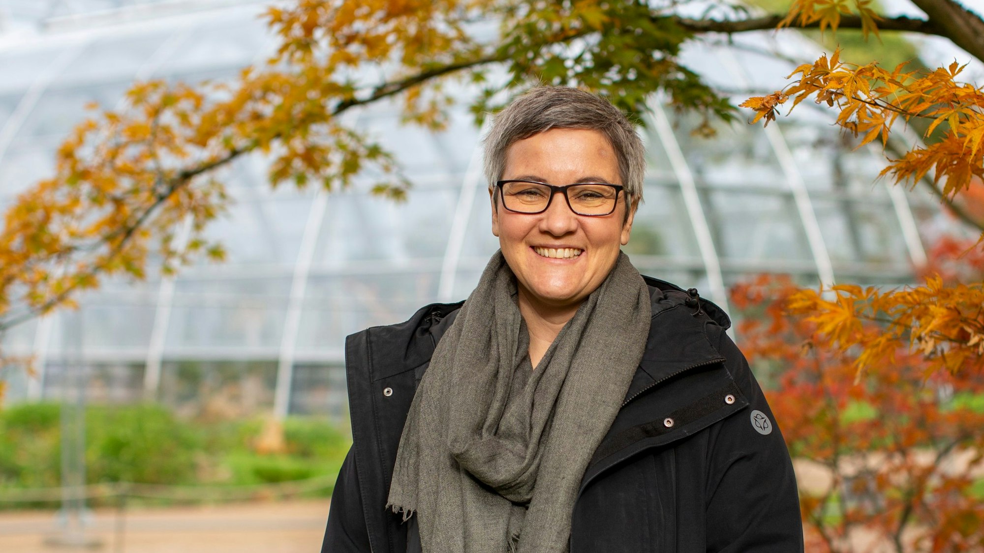 Sie steht unter einem zierlichen Baum mit orangenem Laub, im Hintergrund sind die Schaugewächshäuser zu sehen.
