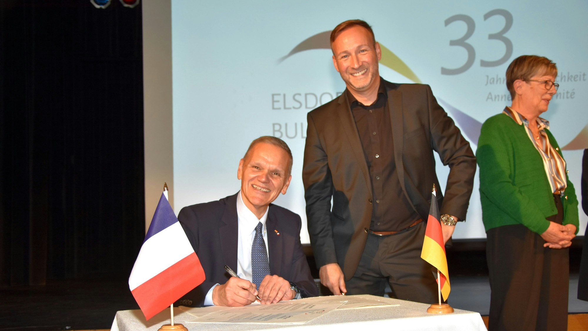 Die Bürgermeister lächeln in die Kamera, einer sitzt an einem Tisch und ist im Begriff, ein Schriftstück zu unterzeichnen.