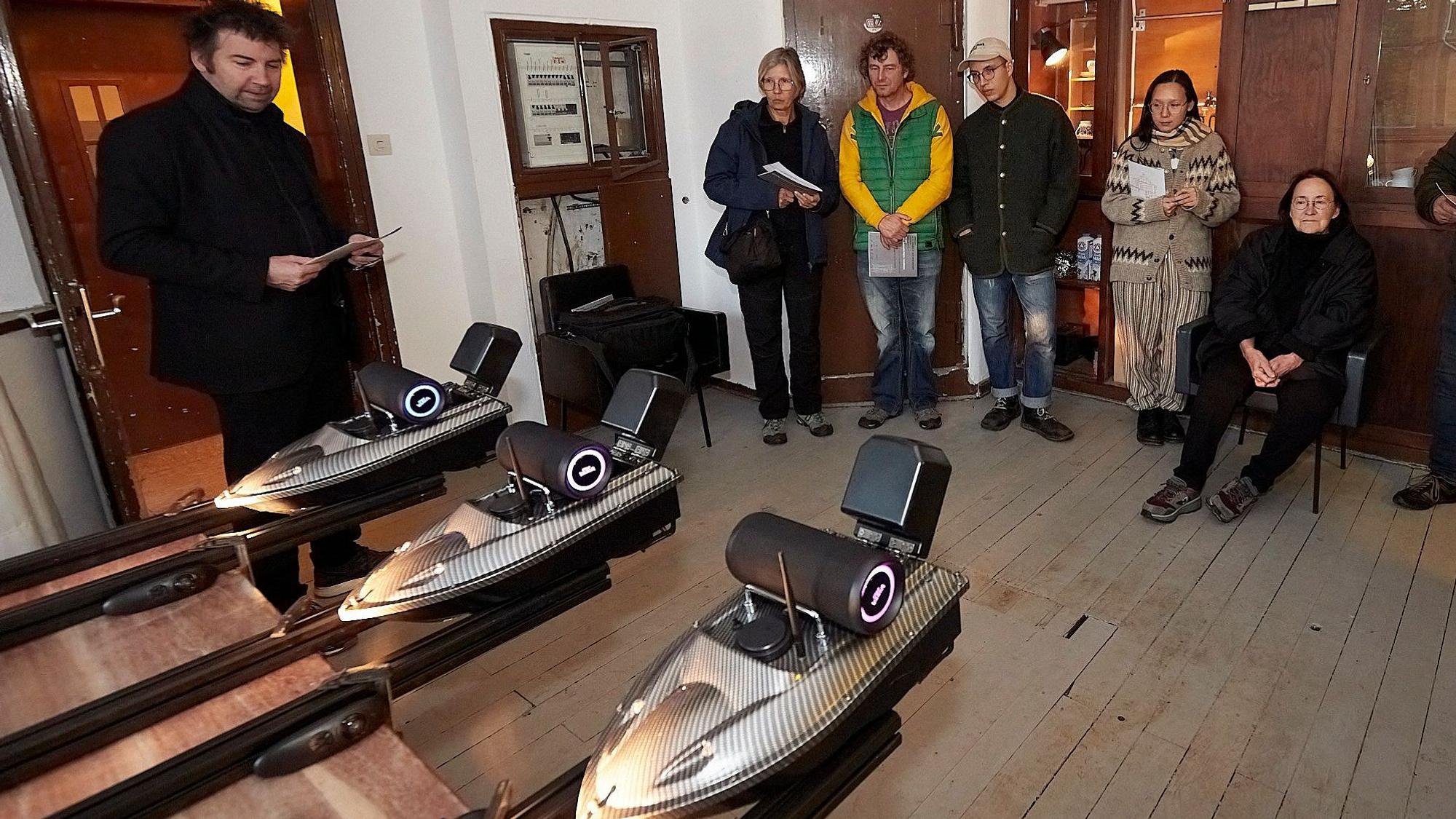 Musikperformance im Bahnhof Scheven: Drei ferngesteuerte Motorboote tragen Lautsprecherboxen, im Hintergrund sind einige Besucher zu sehen.