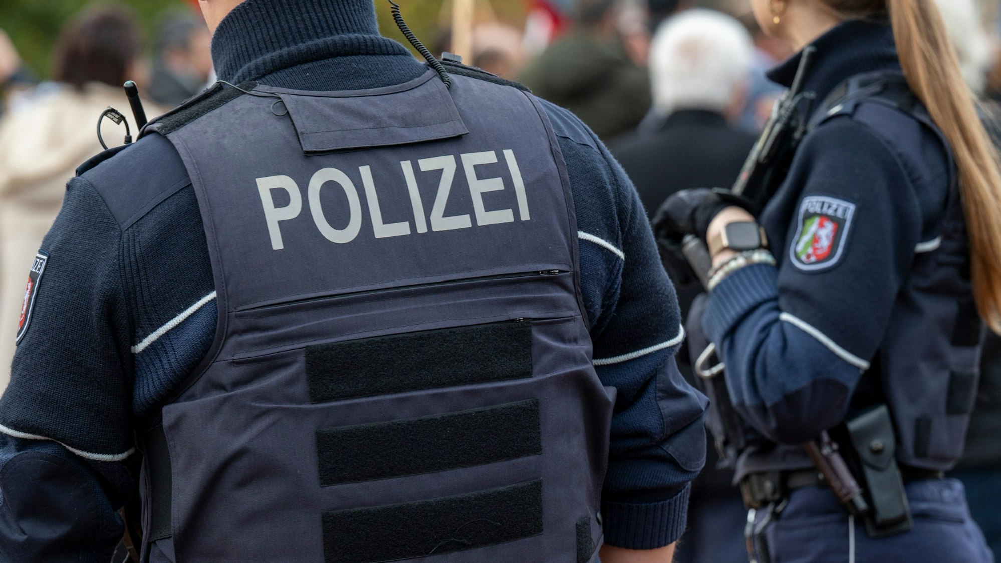 Ein Polizist steht mit dem Rücken zur Kamera. Auf seiner Weste steht groß „POLIZEI“. Neben ihm steht eine Polizistin.