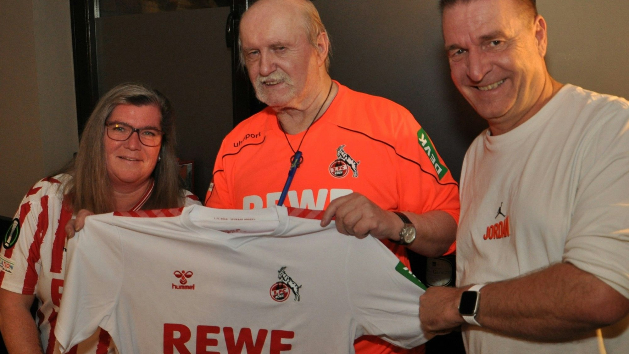 Der FC-Fanbeauftragte Rainer Mendel steht rechts neben Klubgründer Günter Henk daneben steht eine Frau, alle drei alten ein Trikot des 1. FC Köln in die Kamera.
