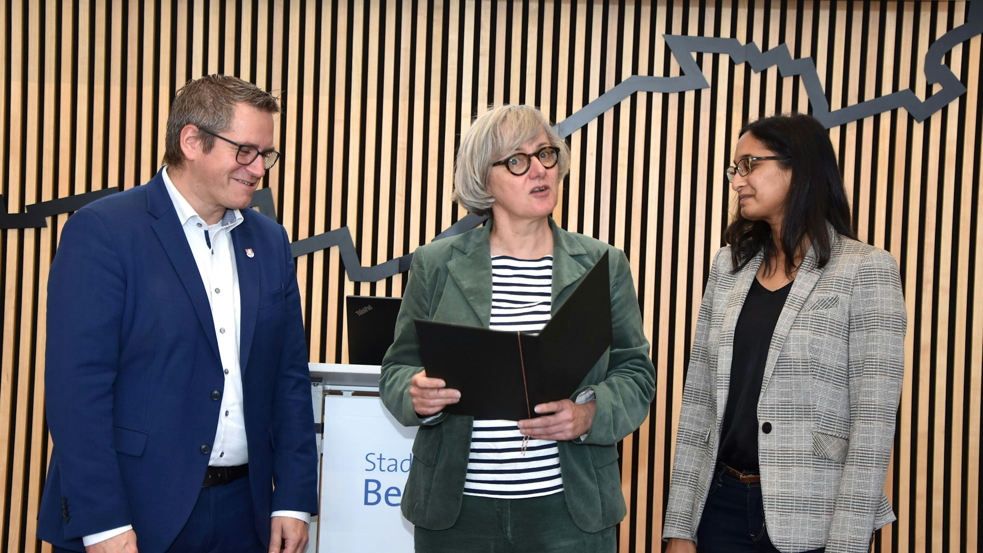 Ein Mann und zwei Frau stehen vor einer holzvertäfelten Wand, die Frau in der Mitte hält eine Kladde.