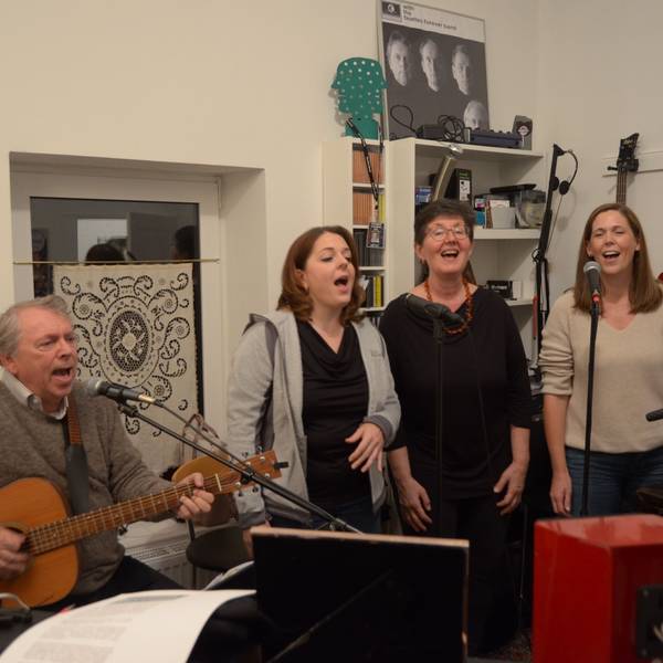 Vier Mitglieder der Band Lenny Pane proben ihr Programm mit Beatles-Liedern. Der Sänger und Gitarrist Manfred Hansen sitzt an einem Mikrofon, neben ihm stehen die Sängerinnen Franziska Lörch, Elise Kushner und Claudia Milz. Im Hintergrund hängen Gitarren und Plakate an der Wand.