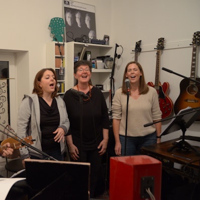 Vier Mitglieder der Band Lenny Pane proben ihr Programm mit Beatles-Liedern. Der Sänger und Gitarrist Manfred Hansen sitzt an einem Mikrofon, neben ihm stehen die Sängerinnen Franziska Lörch, Elise Kushner und Claudia Milz. Im Hintergrund hängen Gitarren und Plakate an der Wand.