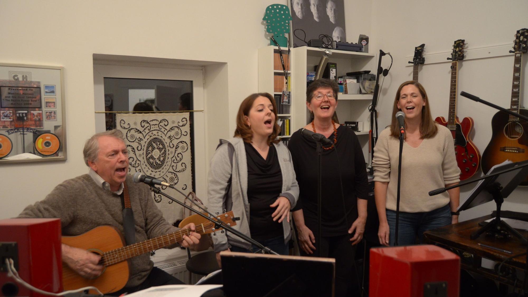 Vier Mitglieder der Band Lenny Pane proben ihr Programm mit Beatles-Liedern. Der Sänger und Gitarrist Manfred Hansen sitzt an einem Mikrofon, neben ihm stehen die Sängerinnen Franziska Lörch, Elise Kushner und Claudia Milz. Im Hintergrund hängen Gitarren und Plakate an der Wand.