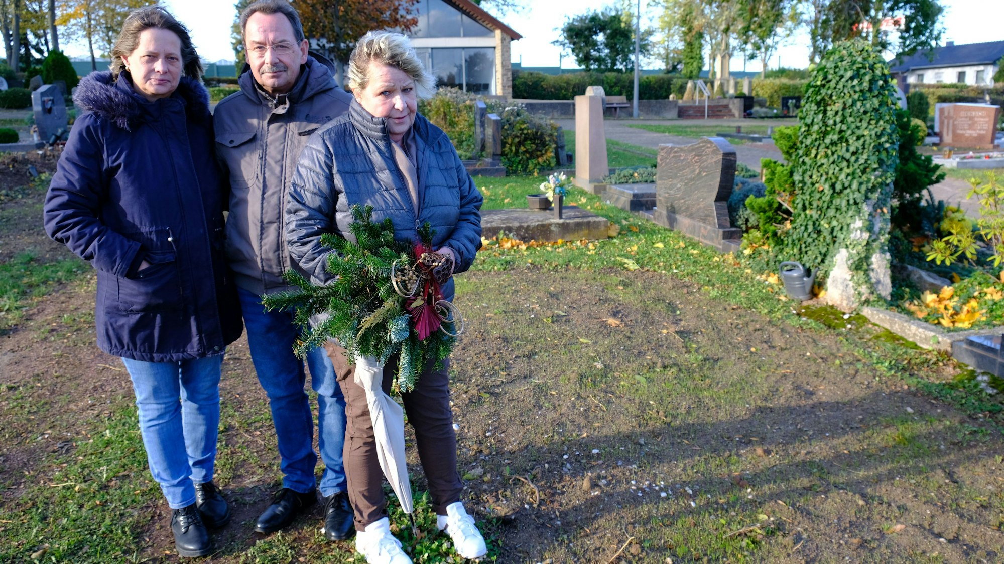 Gabi Große, Bernhard Hoffmann und Anja Hoffmann stehen auf dem Friedhof in Kommern an der Stelle, wo sich bis vor Kurzem noch das Grab ihrer Schwester Sabine befand. Gabi Große hält ein Grabgesteck in der Hand.