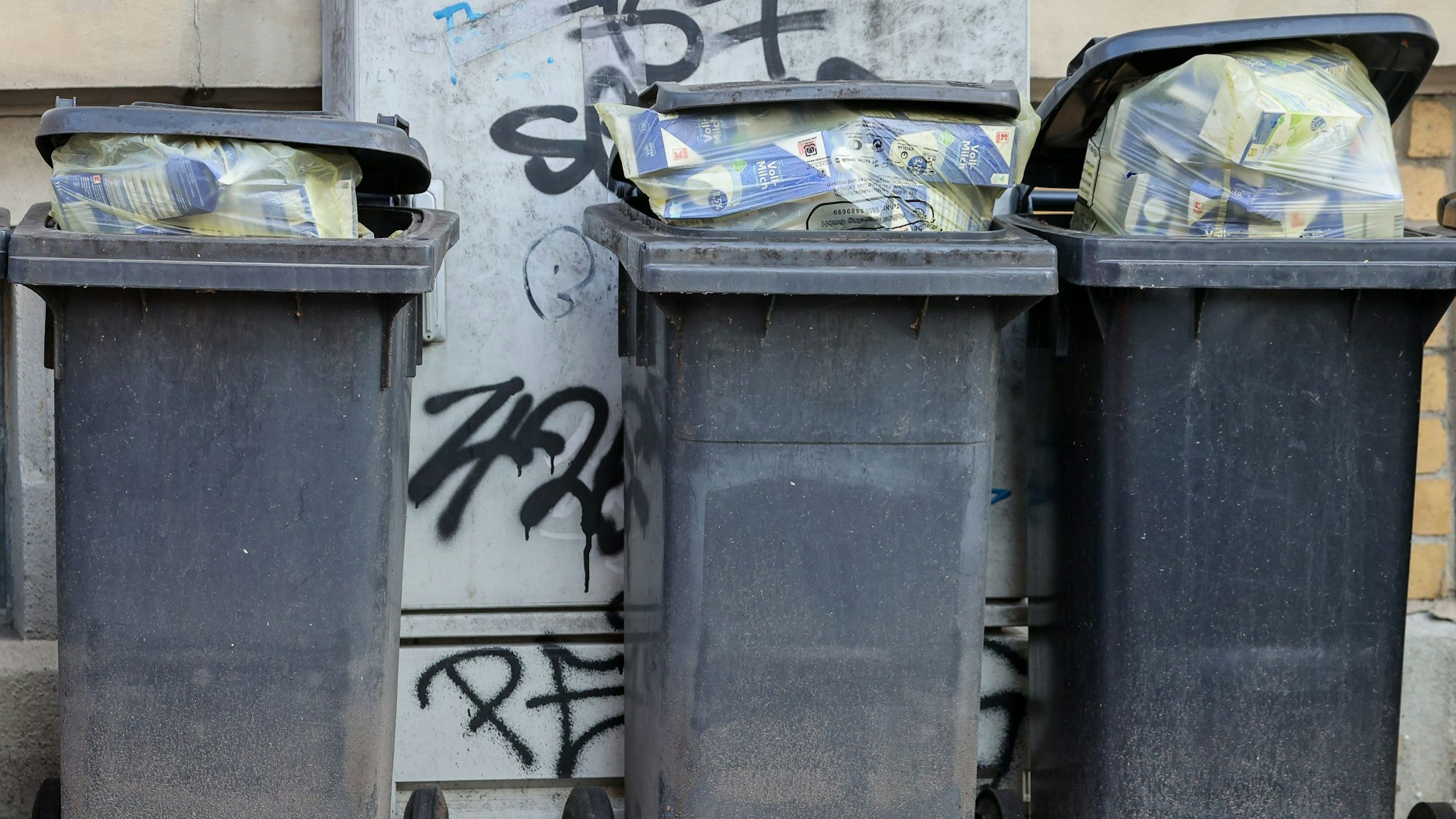 Mehrere volle Mülltonnen für Restmüll stehen an einer Hauswand.