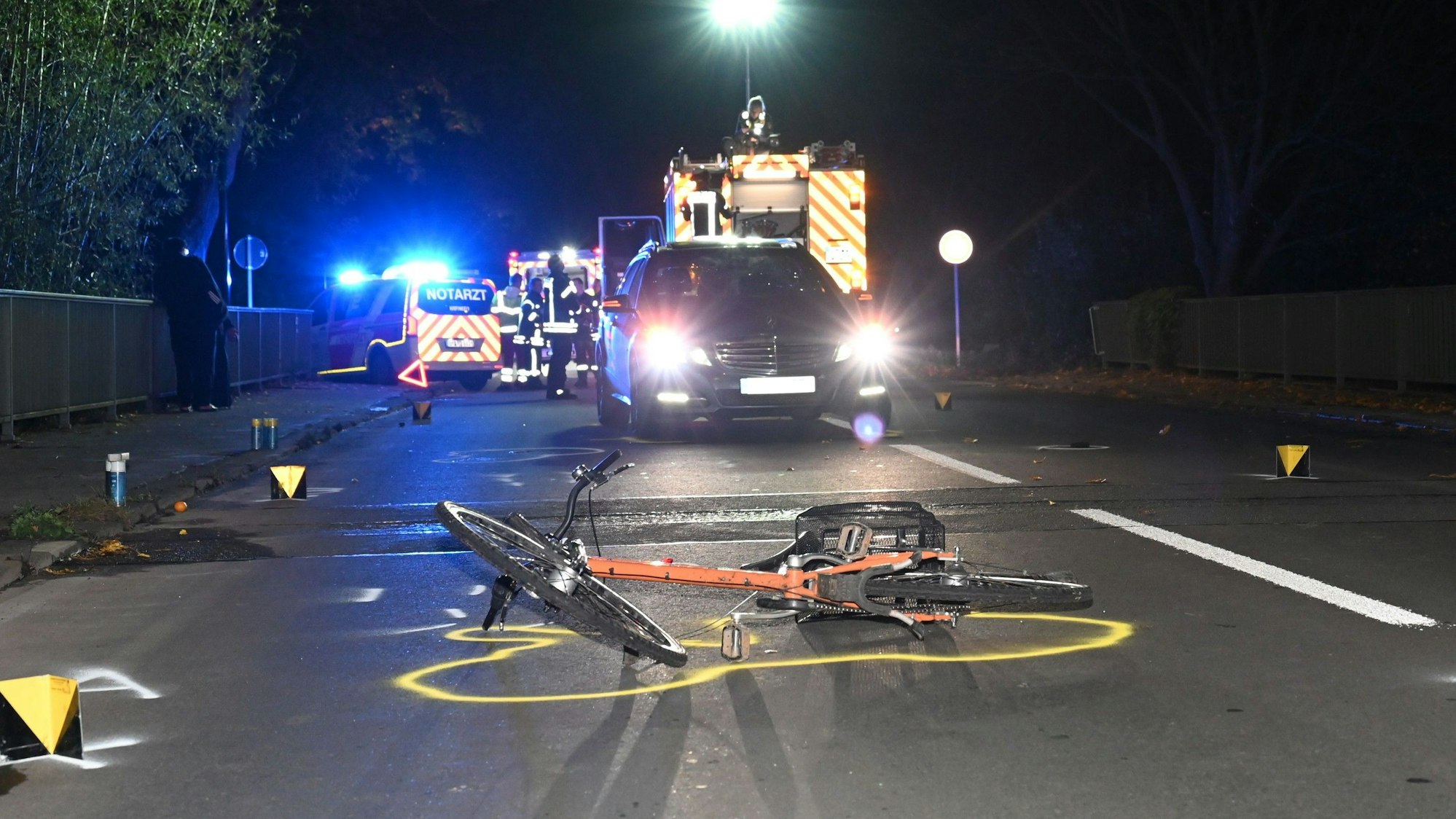 Ein Fahrrad liegt vor einem Pkw und Rettungsfahrzeugen auf einer Straße. Drumherum Markierungen der Polizei.