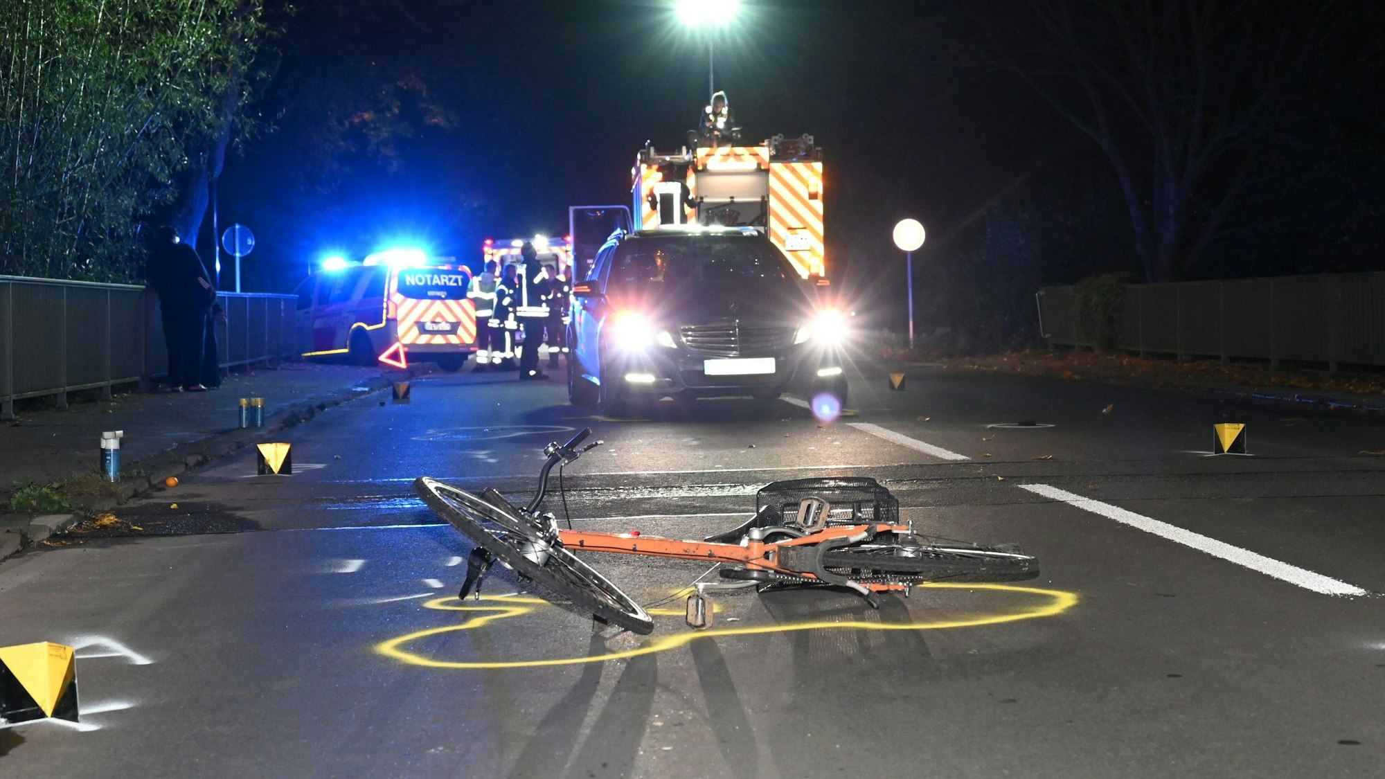 Ein Fahrrad liegt vor einem Pkw und Rettungsfahrzeugen auf einer Straße. Drumherum Markierungen der Polizei.