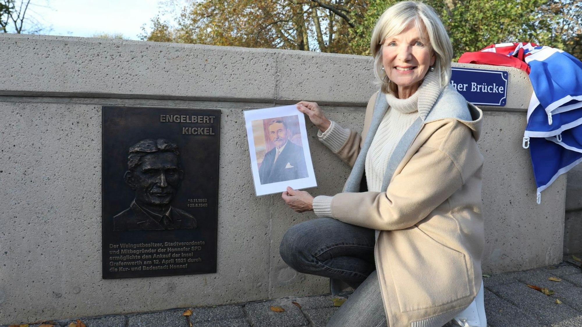 Eine Frau kniet neben einem Relief, das an Engelbert Kickel erinnert. In der Hand hält sie das Bild eines Gemäldes, das Kickel zeigt.