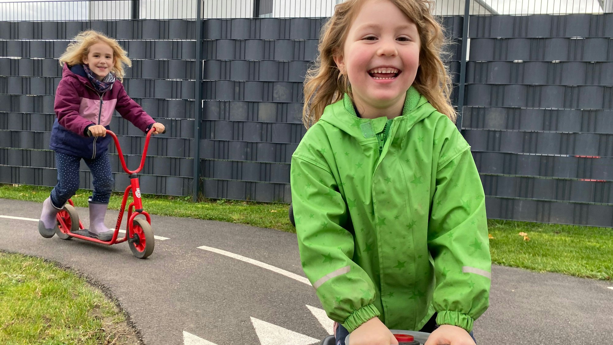 Ein Mädchen auf einem Bobbycar und eines auf einem Roller, fahren schnell in eine Kurve.