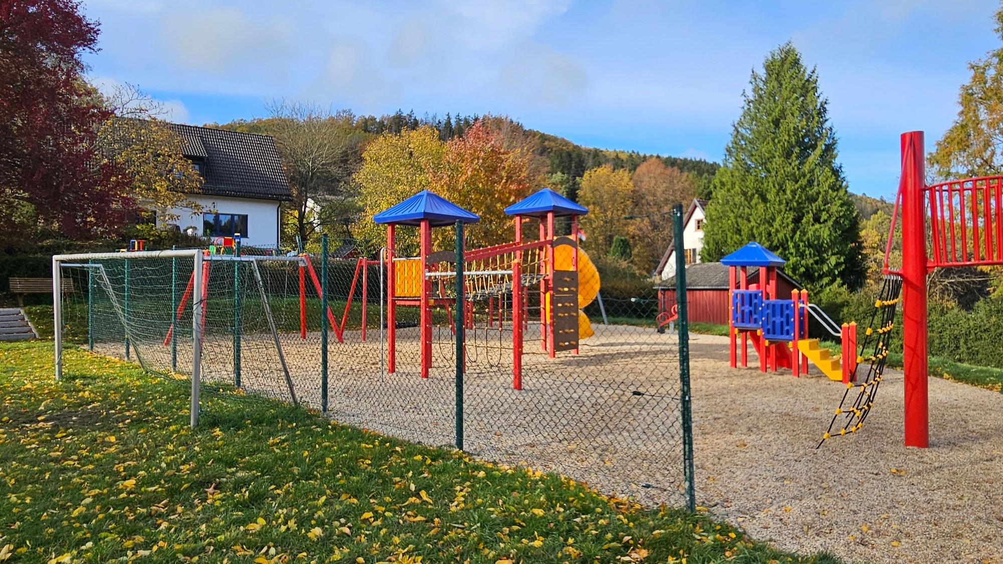 Zu sehen ist das Tor des Bolzplatzes und mehrere bunte Geräte des Spielplatzes.