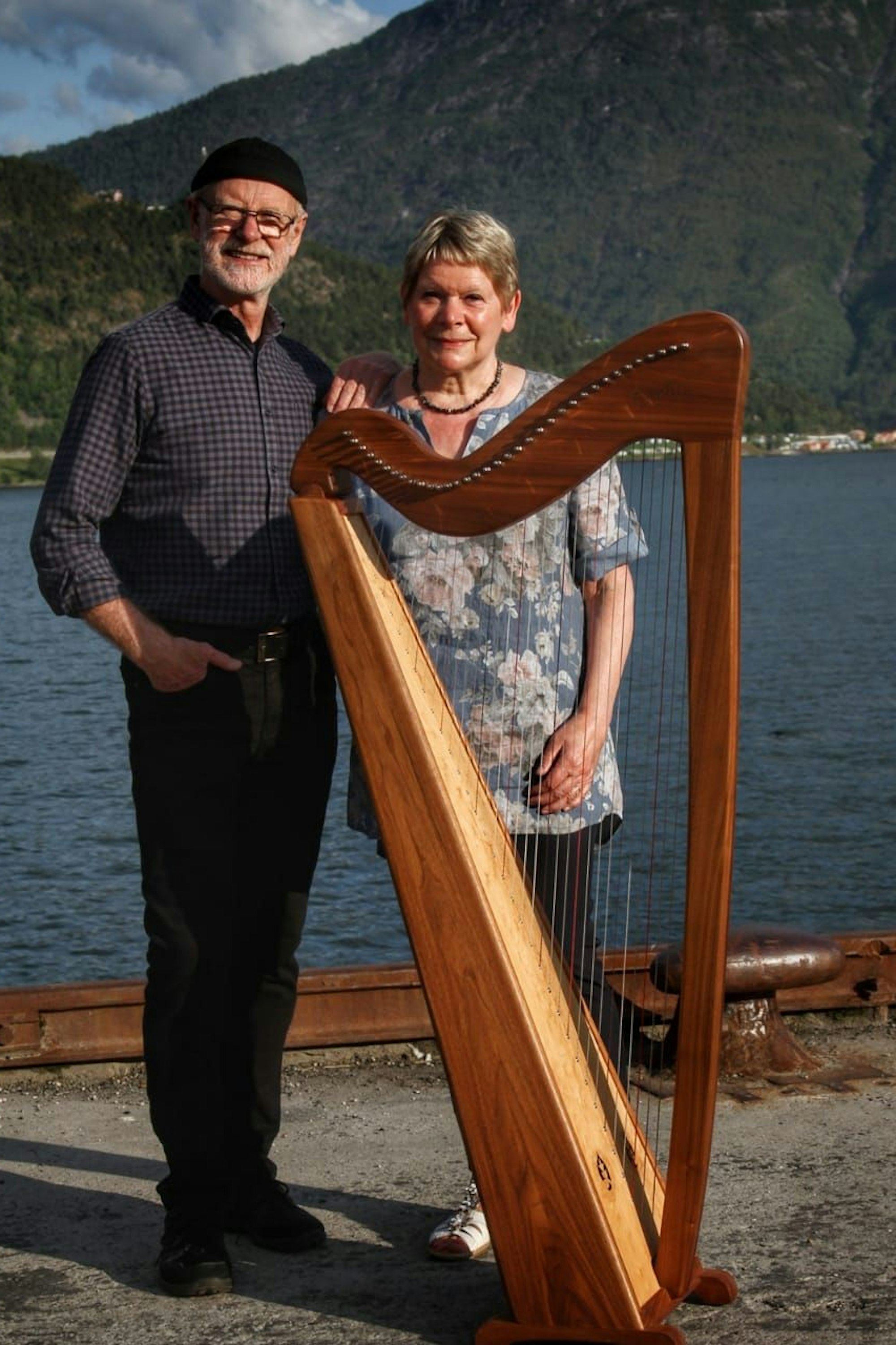 Die beiden Musiker Trude und Leif Johannessen stehen mit einer Konzertharfe am Ufer eines Gewässers.