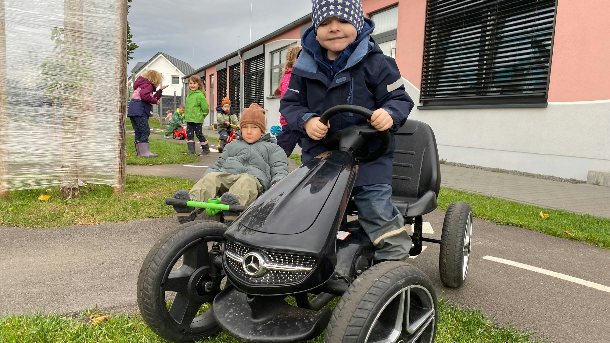 Ein Junge fährt auf einem Kettcar. Es hat eine Kühlerhaube wie ein Mercedes und dicke Reifen.