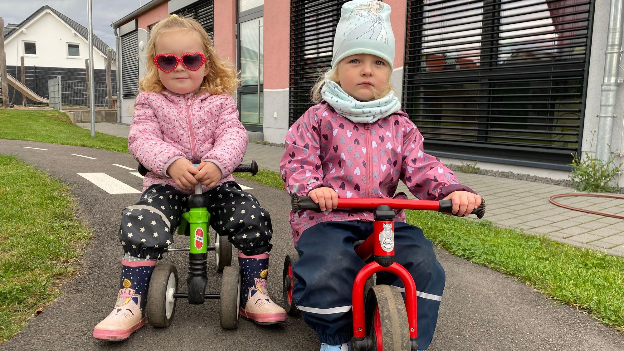 Zwei Mädchen sitzen auf Dreirädern. Eines trägt eine Sonnenbrille.