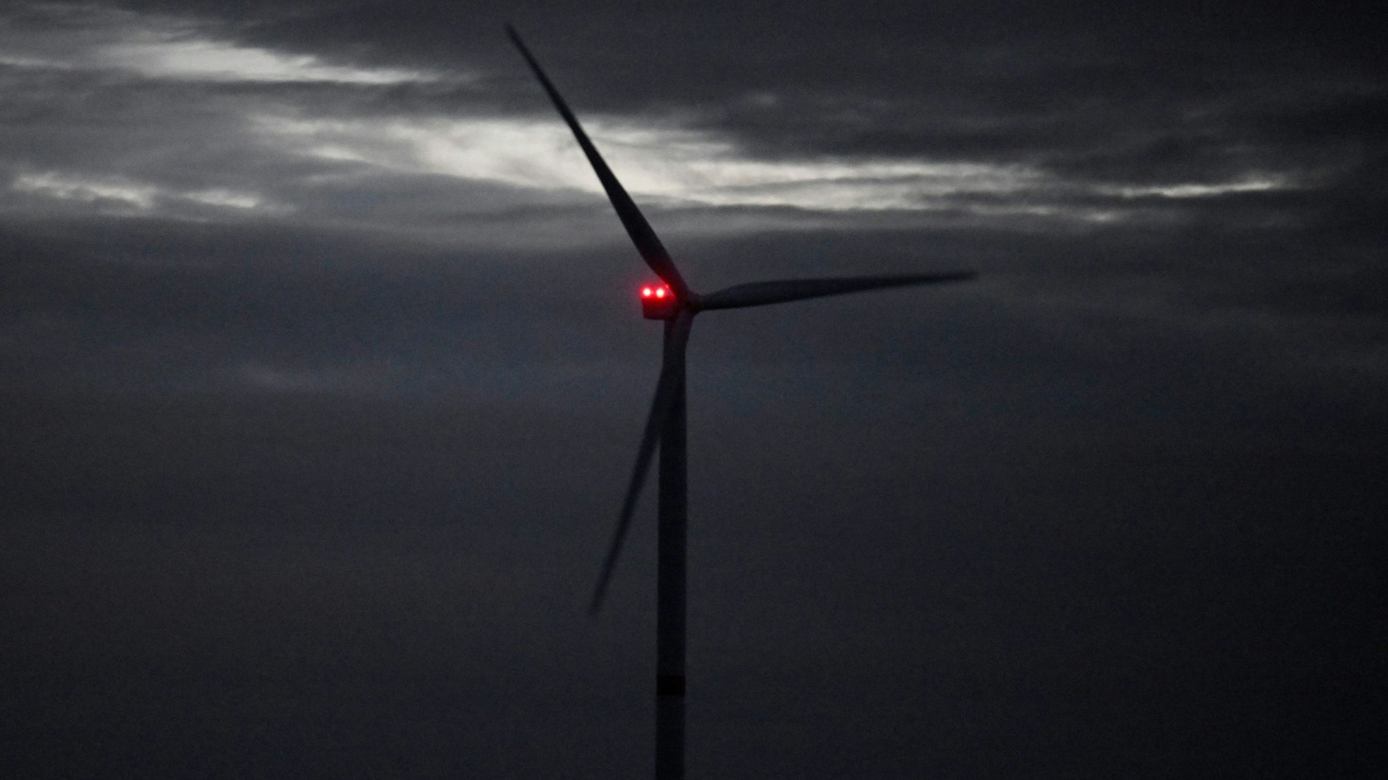 Ein Windrad dreht sich vor einem Unwetter im Wind. Hinter seinen Rotorblättern ist eine rote Leuchte zu sehen.