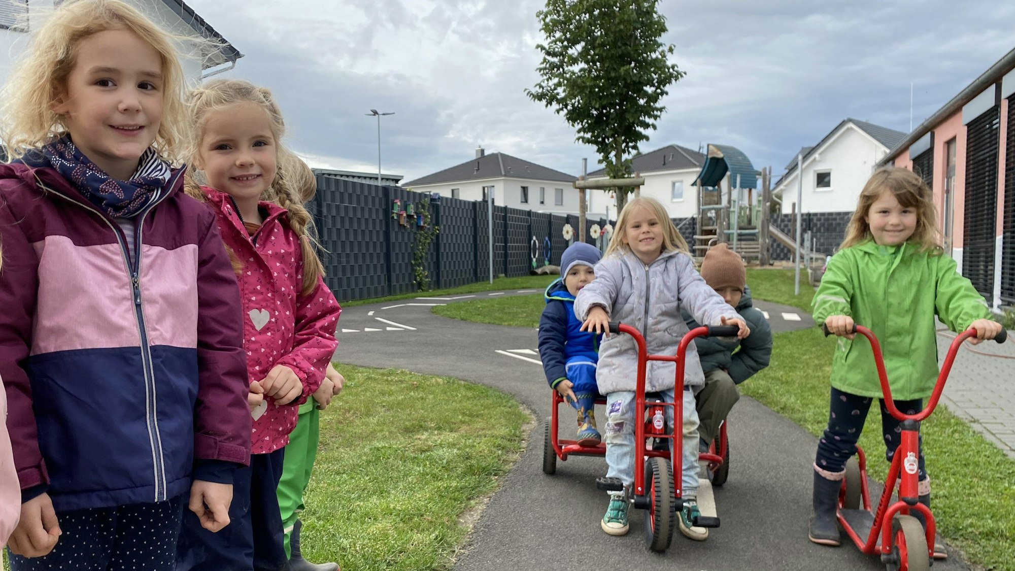 Zwei Mädchen stehen am Straßenrand. Eine Dreiradfahrerin und eine Rollerfahrerin halten auf der schmalen Straße.