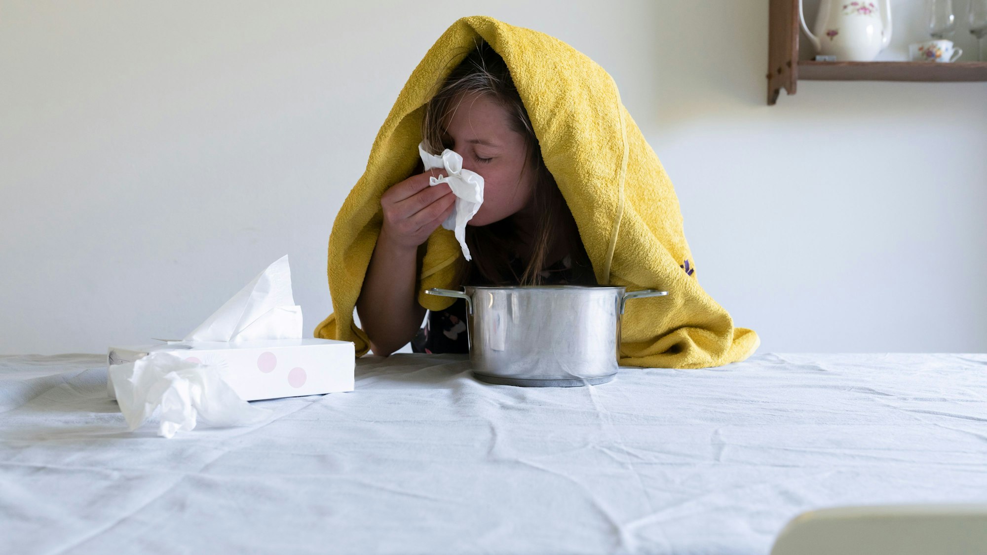Frau mit Handtuch über dem Kopf sitzt vor einem Kochtopf am Tisch und putzt sich die Nase.