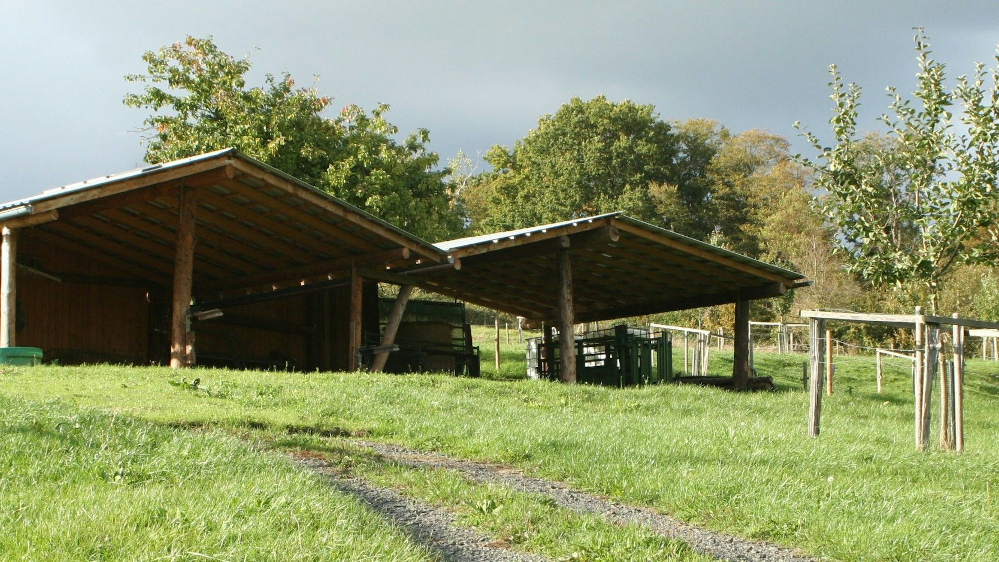 Zwei überdachte, hölzerne Unterstände stehen auf einer Wiese, ein junger Obstbaum steht davor.