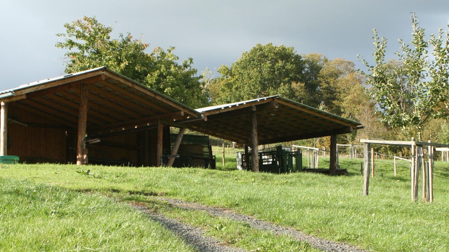 Zwei überdachte, hölzerne Unterstände stehen auf einer Wiese, ein junger Obstbaum steht davor.