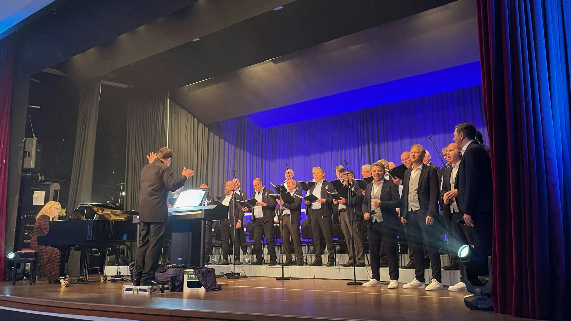 Auf einer Bühne, eingetaucht in blaues Licht, stehen Männer im Anzug vor Mikrofonen und singen. Mit ihnen auf der Bühne sind ein Dirigent und eine Pianistin am Flügel.