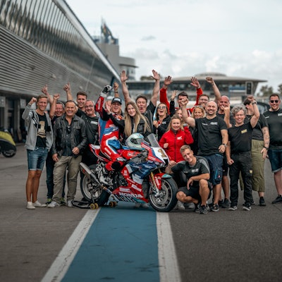 Eine Gruppe steht jubelnd auf einer Rennstrecke. In der Mitte von ihnen ist der oberbergischen Motorradrennfahrer Florian Alt auf seiner Honda.