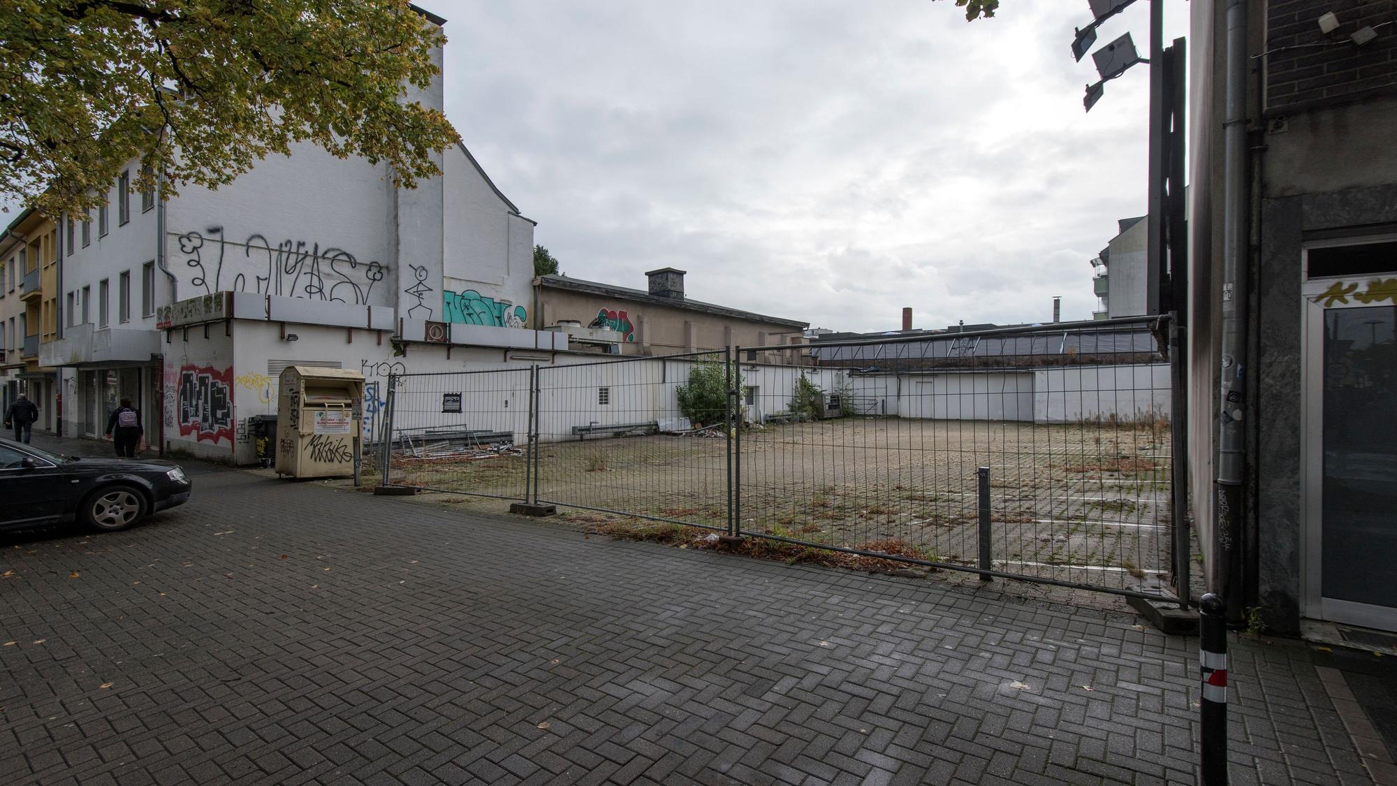Hinter einem Bauzaun liegt eine Brachfläche, links und rechts sind Häuser.