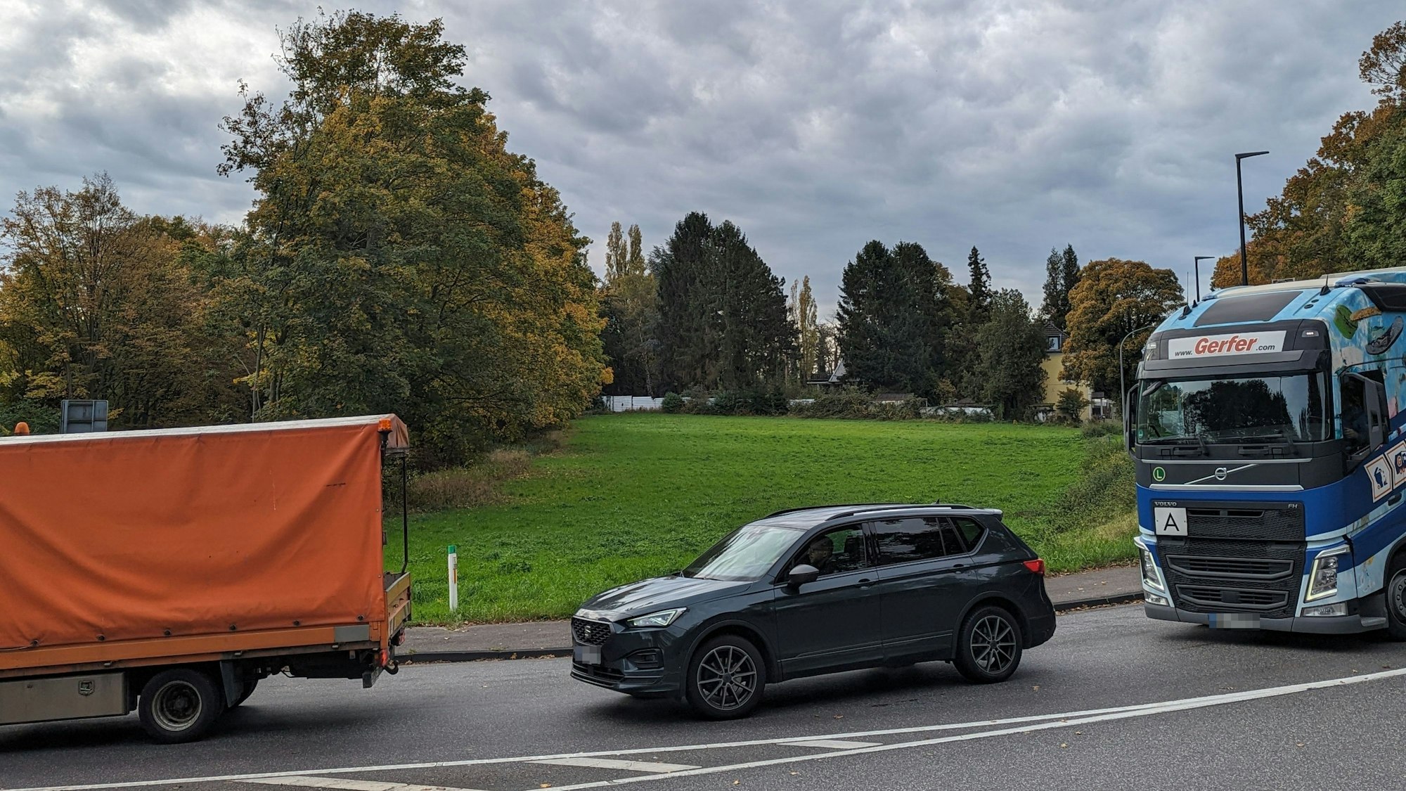 Autos und Transporter fahren an einer Wiese über eine stark befahrene Straße.