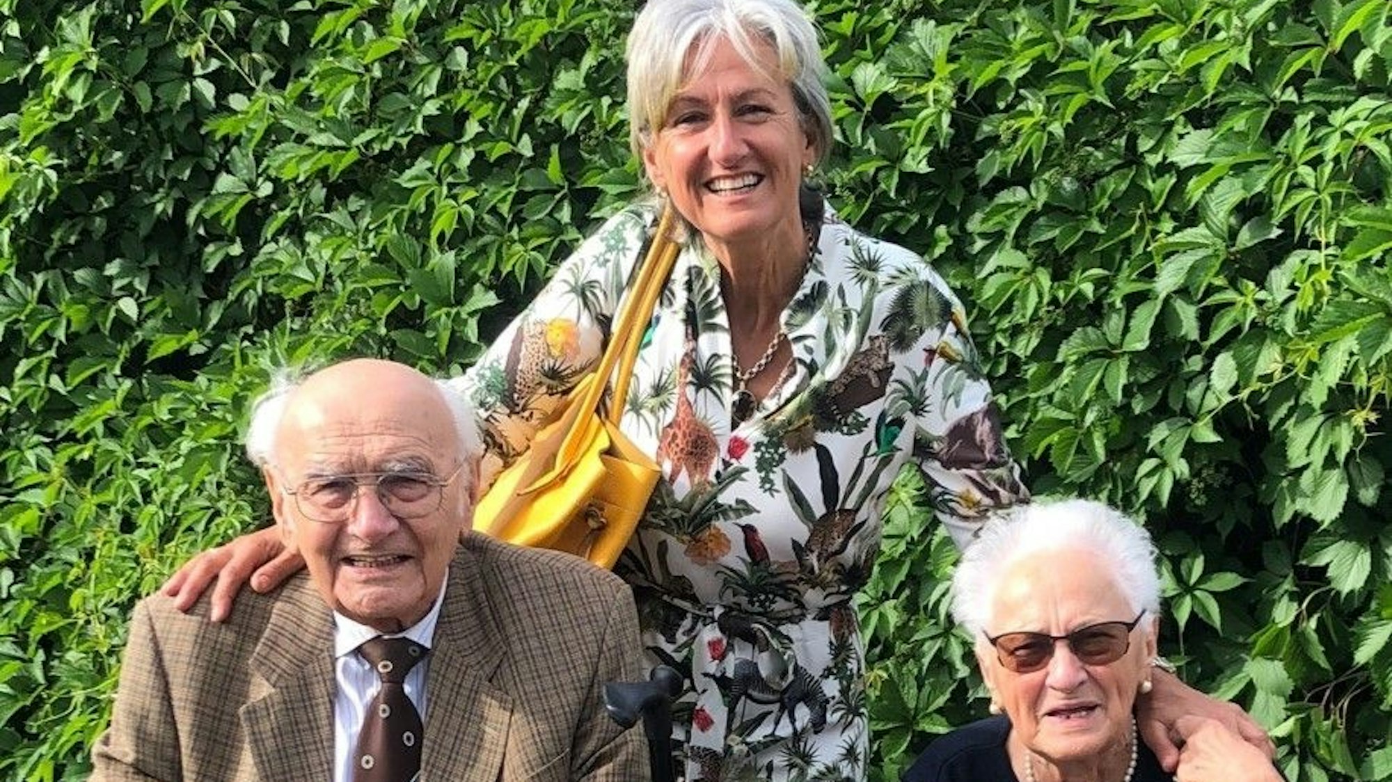 Eine Frau steht zwischen einem älteren Mann und einer älteren Frau und legt ihren Hände auf die Schultern der beiden.