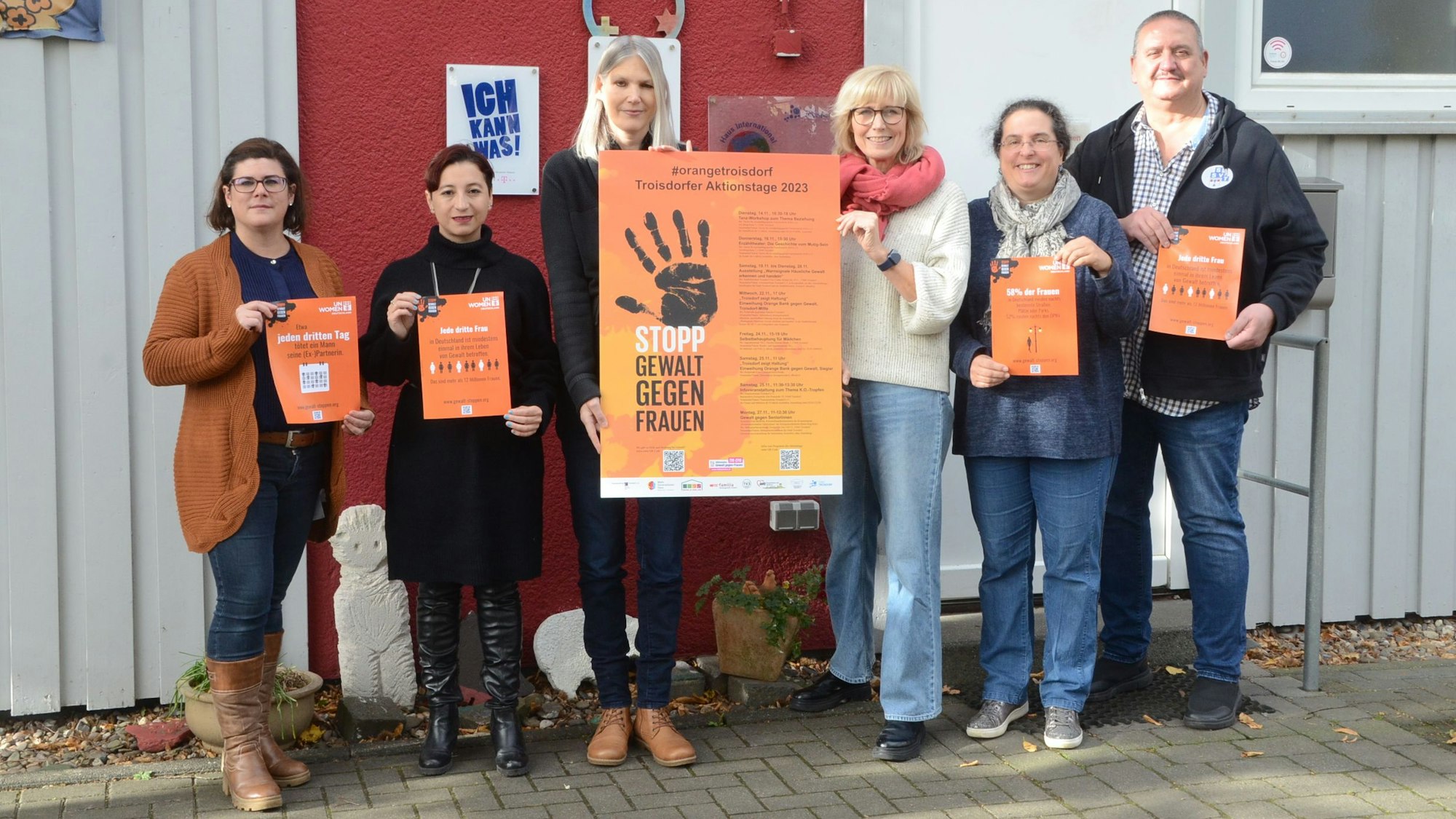 Fünf Frauen und ein Mann stehen vor einer Hauswand in Grau und Dunkelrot. Sie halten Plakate in der Hand. Auf einem großen Plakat sind eine schwarze Hand und der Satz Stopp Gewalt Gegen Frauen zu sehen.