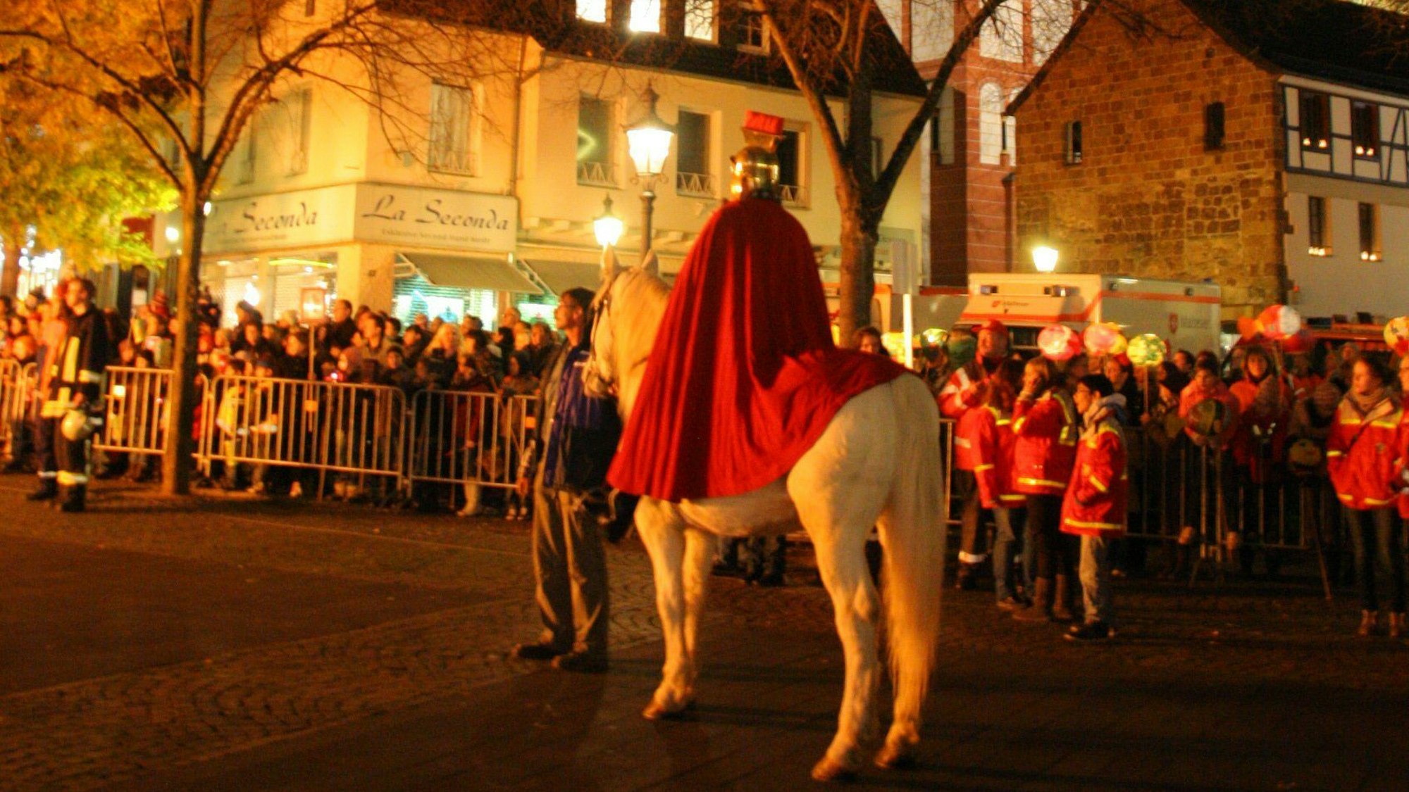 Ein Mann im Kostüm eines römischen Legionärs sitzt auf einem weißen Pferd. Im Hintergrund stehen viele Menschen, vor allem Kinder. Viele tragen Martinslaternen.