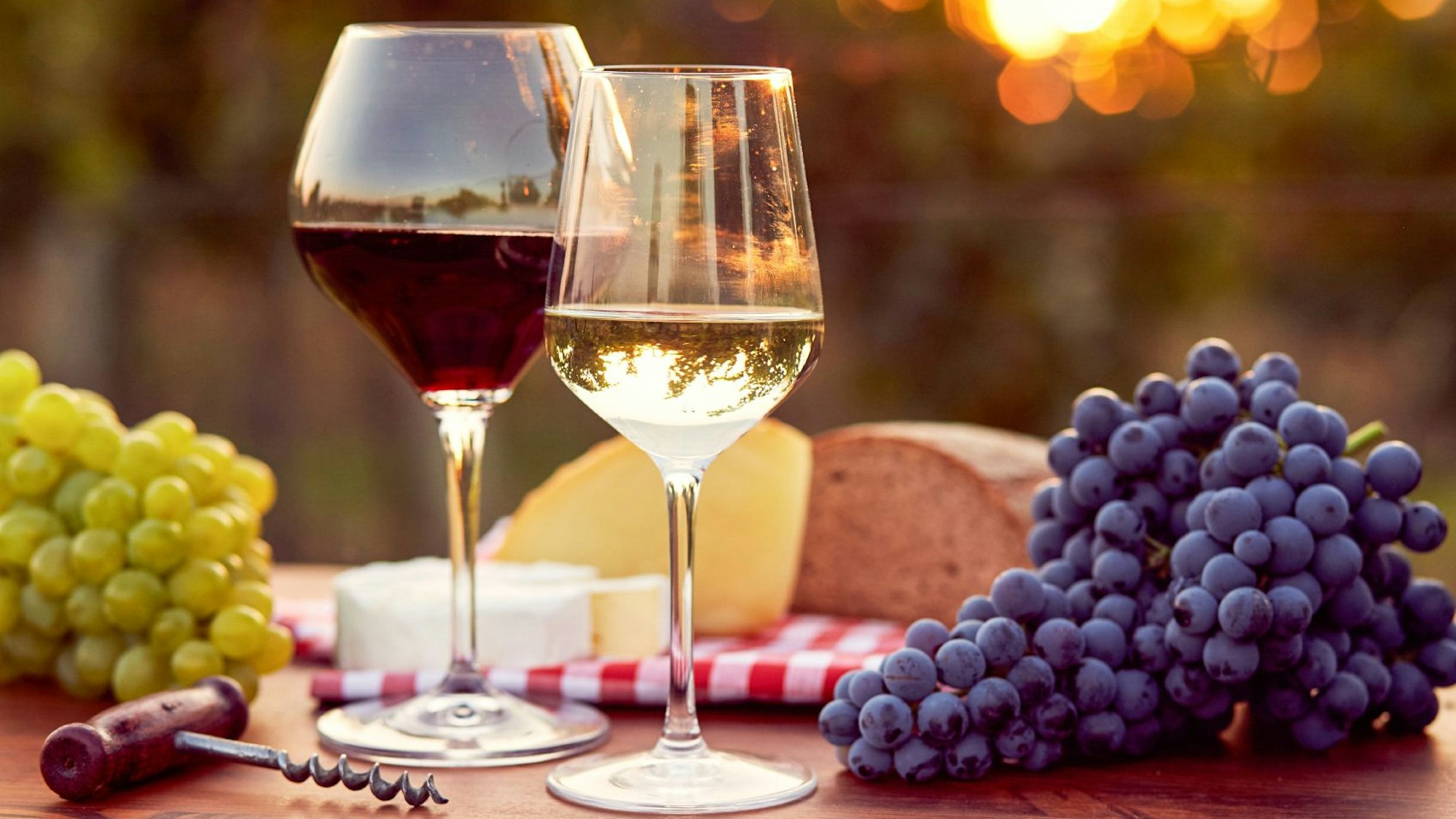 Ein Tisch mit zwei Gläsern Wein, Brot, Käse, Trauben und einem Korkenzieher.