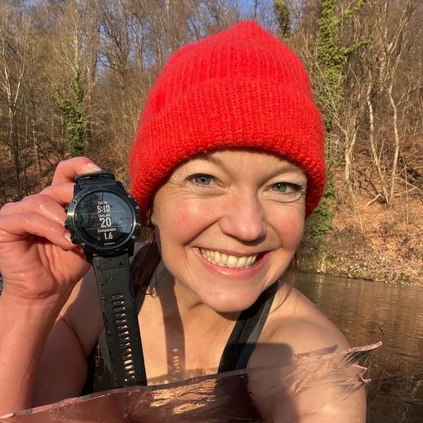 Anita Horn sitzt am Aggerufer in Schwimmkleidung und einer roten Wollmütze. Sie zeigt eine Armbanduhr mit Temperaturanzeige.