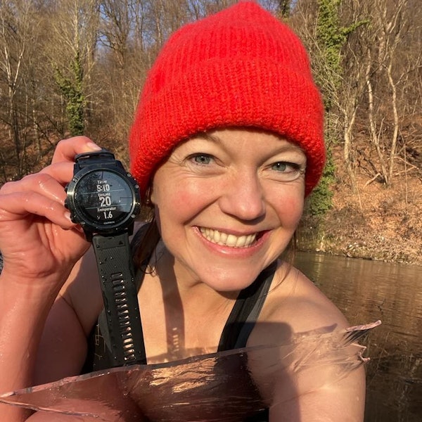 Anita Horn sitzt am Aggerufer in Schwimmkleidung und einer roten Wollmütze. Sie zeigt eine Armbanduhr mit Temperaturanzeige.