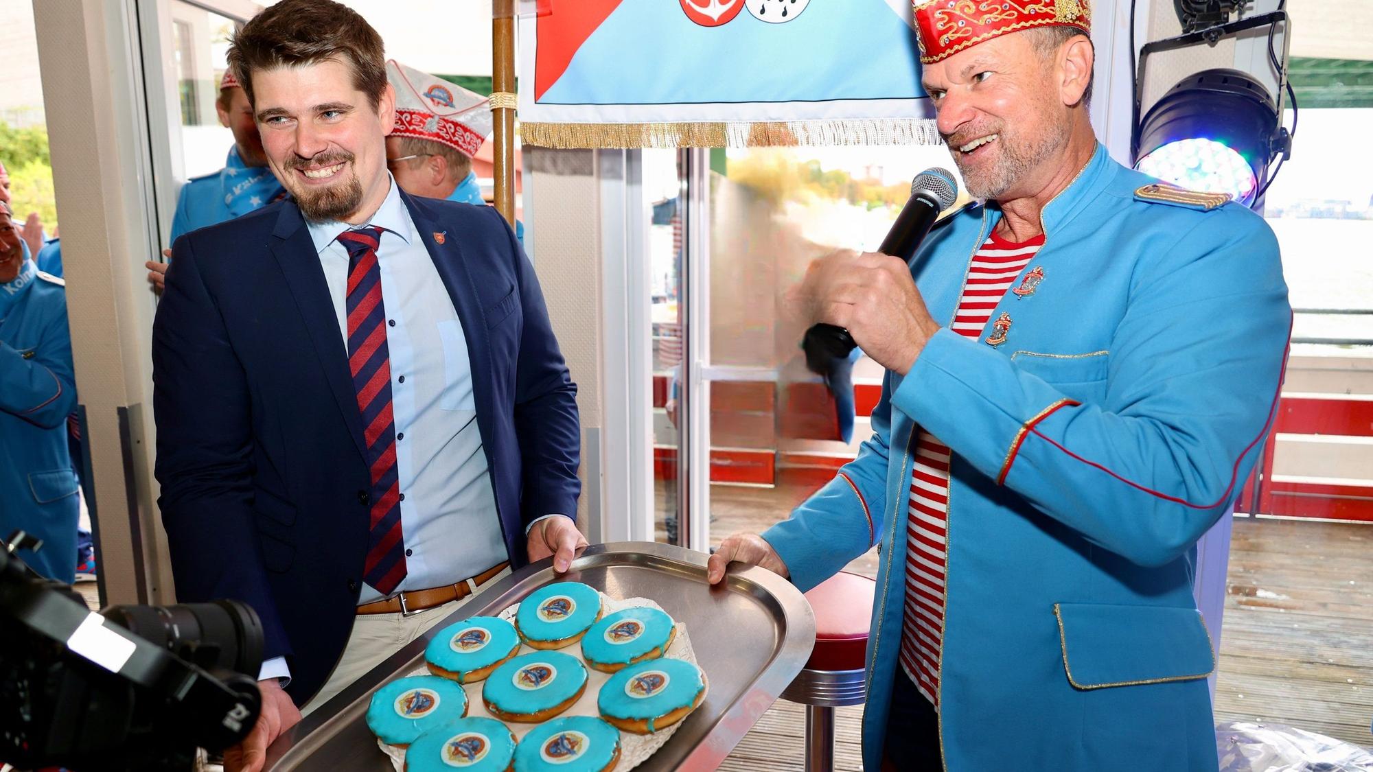 Ein Mann im Anzug und einer mit hellblauem Frack, Ringel-Shirt und Karnevalsmütze.