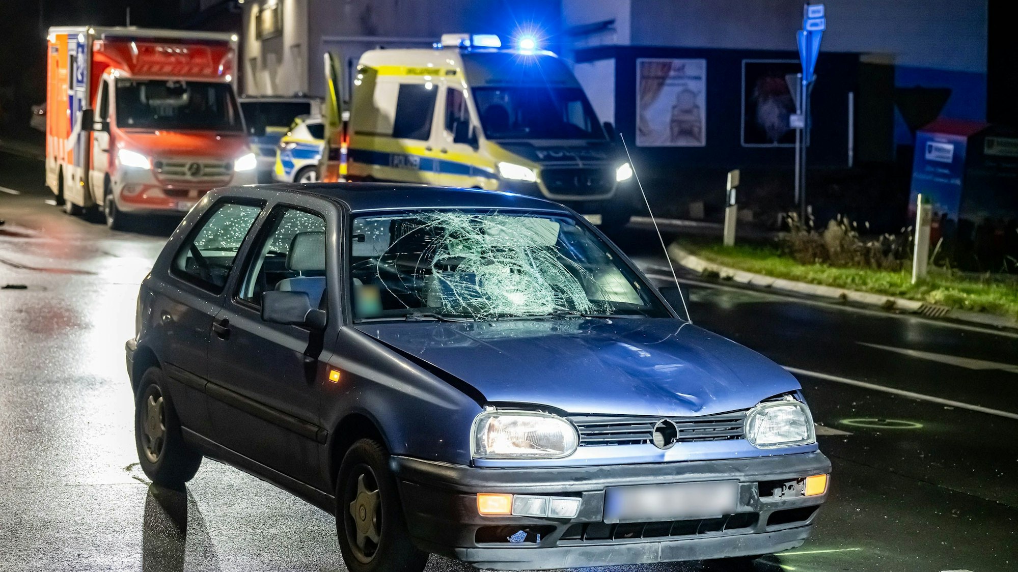 Ein VW-Golf steht mit sichtbaren Unfallspuren auf einer Straße, auf dem Boden sind aufgesprühte Markungen zur Unfallaufnahme zu sehen. Im Hintergrund Einsatzfahrzeuge von Polizei und Rettungsdienst mit eingeschaltetem Blaulicht.