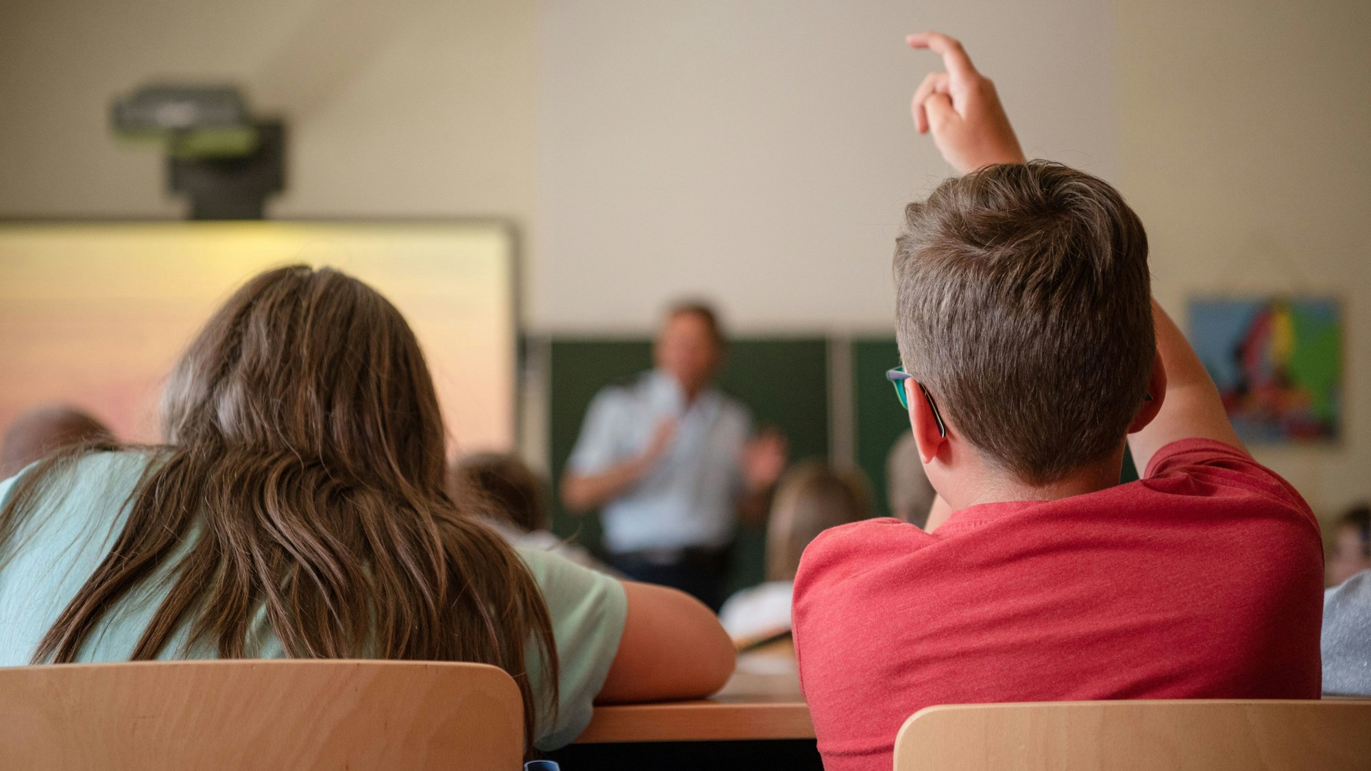 Ein Junge und ein Mädchen sind von hinten zu sehen. Sie sitzen in einer Schulklasse. Der Junge meldet sich.